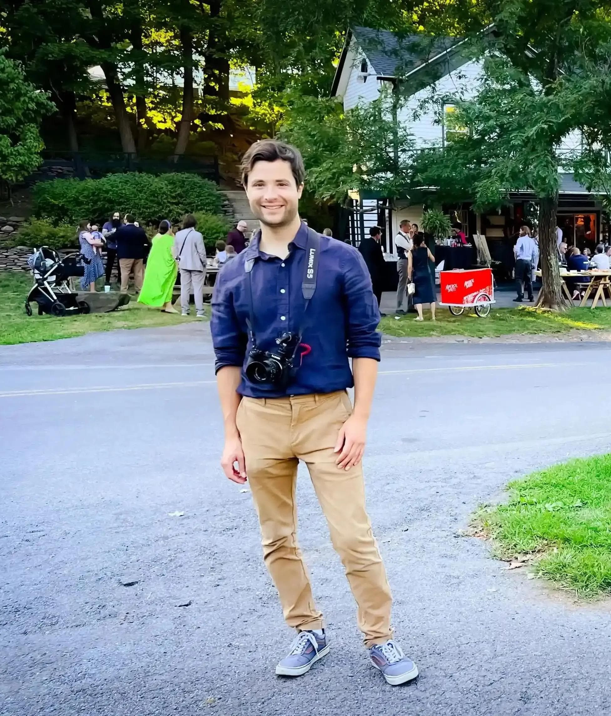 Noah Dickinson, founder of NoahFest Weddings, smiling outdoors at a wedding venue with a camera around his neck."